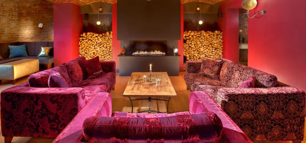 A cozy lounge with plush red and purple sofas around a wooden coffee table, a modern fireplace, stacked firewood, warm lighting, and exposed brick walls creating an inviting atmosphere.