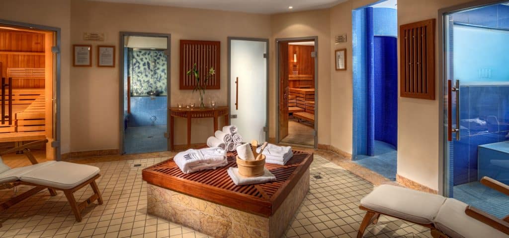 A spa room with wooden benches, rolled towels, a bucket, multiple saunas, steam rooms, and a blue-tiled shower area. The space features warm lighting and a clean, relaxing atmosphere.