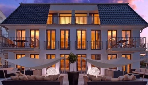 Modern three-story building with large windows and balconies, warmly lit from inside at sunset. Patio has outdoor seating, white shade sails, decorative lighting, and a central potted plant.