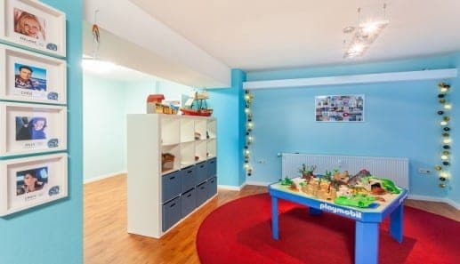 A bright playroom with blue walls, a large playmobil table on a red rug, a white storage unit with blue bins, and framed photos of children on the wall. String lights hang near a radiator.