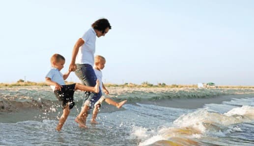 An adult and two young children, all wearing light shirts and shorts, joyfully kick water at the edge of a sandy beach on a bright, sunny day. The adult holds the childrens hands as they play together.