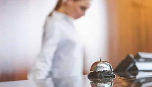 A hotel reception desk with a service bell in focus at the front; a blurred person in professional attire stands in the background.