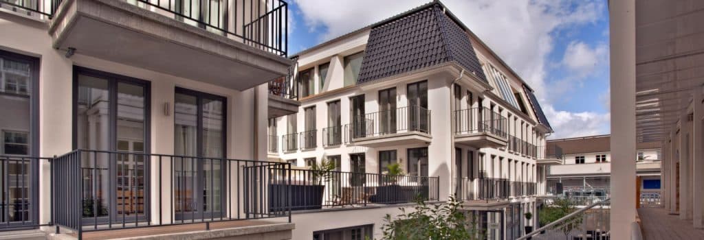 Modern apartment buildings with cream-colored facades, large windows, and metal railings on balconies, set around a central courtyard under a partly cloudy sky.